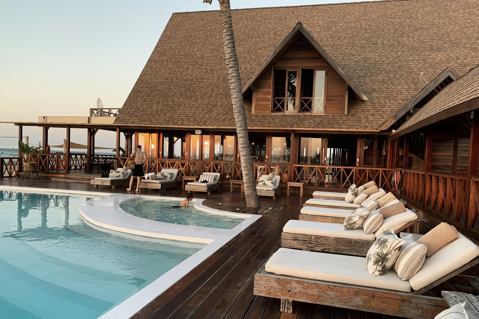 Deck de madeira com espreguiçadeiras à beira da piscina de uma pousada, ao entardecer